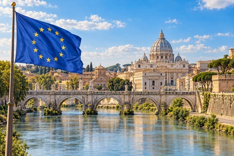 European flag beside a sunlit river and historic European architecture.